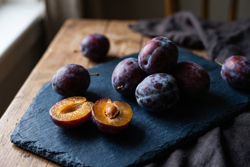 Dark purple plums with a dusty bloom, one halved showing amber flesh, the plum note in perfume