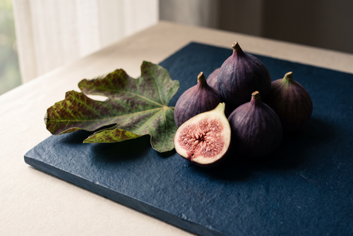 Ripe purple figs, one split open beside a fig leaf on dark slate, the fig note in fragrance