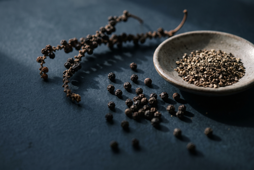 Black peppercorns, a dried pepper vine and a dish of cracked pepper on dark stone, the black pepper note in fragrance