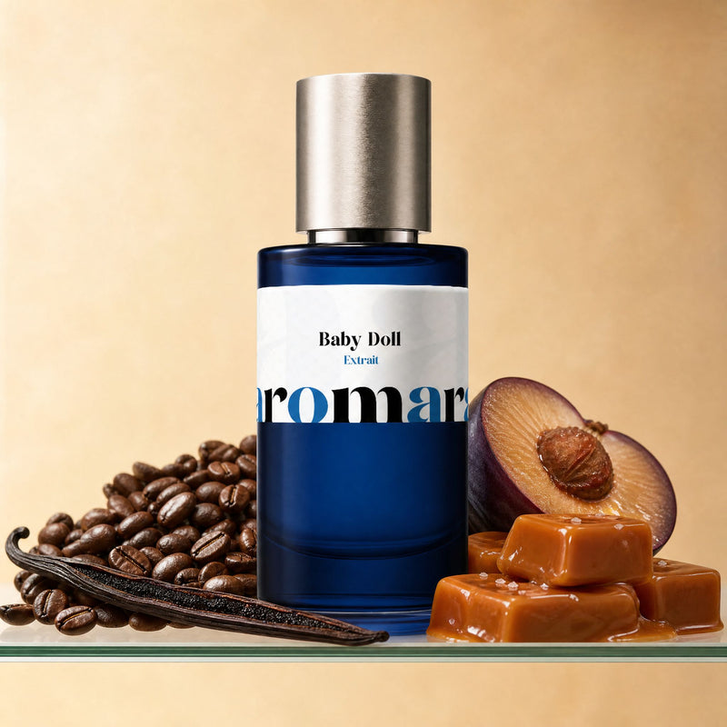 Aromara Baby Doll Extrait perfume bottle with coffee beans, vanilla pod, caramel chunks, and halved plum on a clear glass shelf, warm caramel-cream backdrop