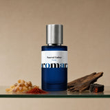 Aromara Sacred Ember Extrait perfume bottle on glass shelf with frankincense resin, saffron threads, oud wood and ember-glow charcoal on soft warm taupe background