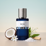 Salt Skin by Aromara on a pale sea-mist blue to soft sand beige background with a halved coconut, a tiare flower, sea salt crystals, lily of the valley and sandalwood. 35% Extrait, inspired by Phlur Beach Skin.