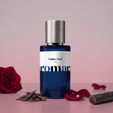 Caden Oud by Aromara on a pale dusty rose grey to soft wine rose background with a deep red rose, oud wood chips, red rose petals and an agarwood log. 35% Extrait, inspired by Oman Luxury Caden.